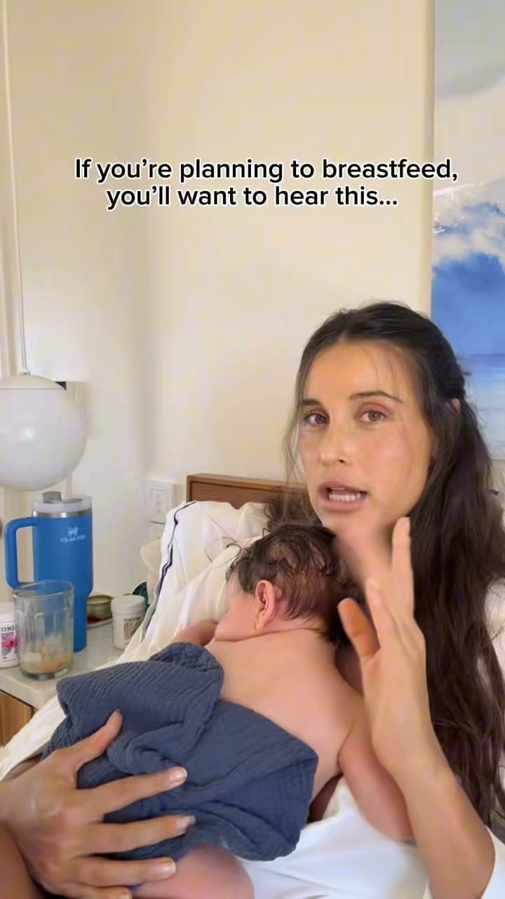 A woman is holding a newborn baby while speaking directly to the camera. On-screen text reads, "If you're planning to breastfeed, you'll want to hear this...". The woman appears to be sharing advice or information related to breastfeeding. The background shows a bedroom setting with a blue insulated mug and other items on a nightstand.