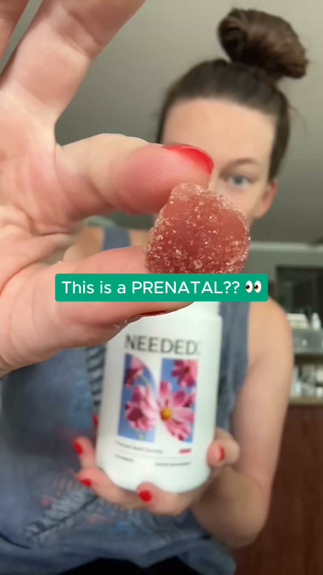 A woman holds up a red gummy in her fingers, showing it to the camera. Behind her, she holds a white bottle of Needed. prenatal gummies. A green text box overlays the image with the question "This is a PRENATAL?? 👀". The woman appears to be a creator filming herself, likely sharing her surprise or opinion about the gummy's appearance or taste, implying it doesn't look like a typical prenatal vitamin.