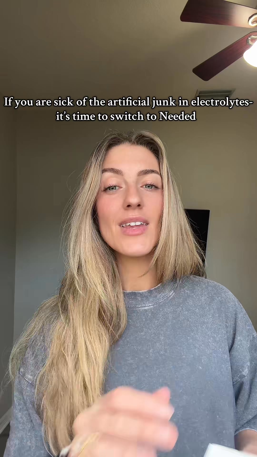 A woman with long blonde hair is looking directly at the camera. She is wearing a grey t-shirt. White text is overlaid on the top of the image. The text reads: "If you are sick of the artificial junk in electrolytes- it's time to switch to Needed". The woman is smiling and appears to be speaking. The background is a plain wall with a ceiling fan visible at the top.