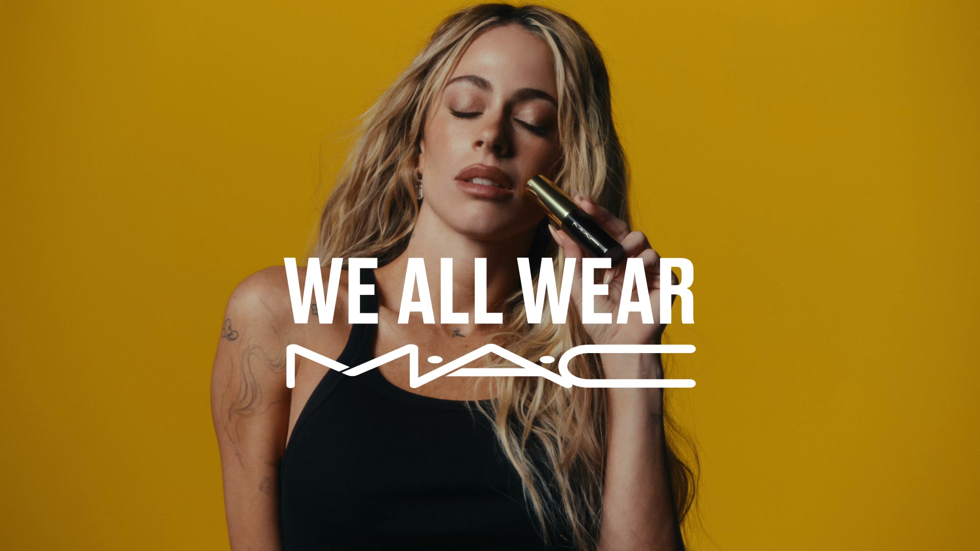 A woman with blonde, wavy hair is shown with her eyes closed, holding a MAC lipstick tube near her lips against a bright yellow background. Large white text overlays the image, reading "WE ALL WEAR" with the MAC logo below it. The woman is wearing a black tank top and has tattoos on her arm. The overall aesthetic is a close-up shot focused on beauty and brand identity.