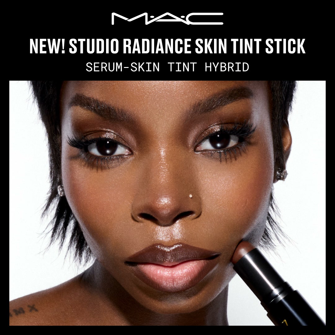 A close-up shot of a woman's face with dark skin and dramatic makeup is shown. She is holding a MAC Studio Radiance Skin Tint Stick up to her cheek. The MAC logo is at the top of the image, with the text "NEW! STUDIO RADIANCE SKIN TINT STICK" and "SERUM-SKIN TINT HYBRID" displayed prominently. The image is a product advertisement for MAC Cosmetics.
