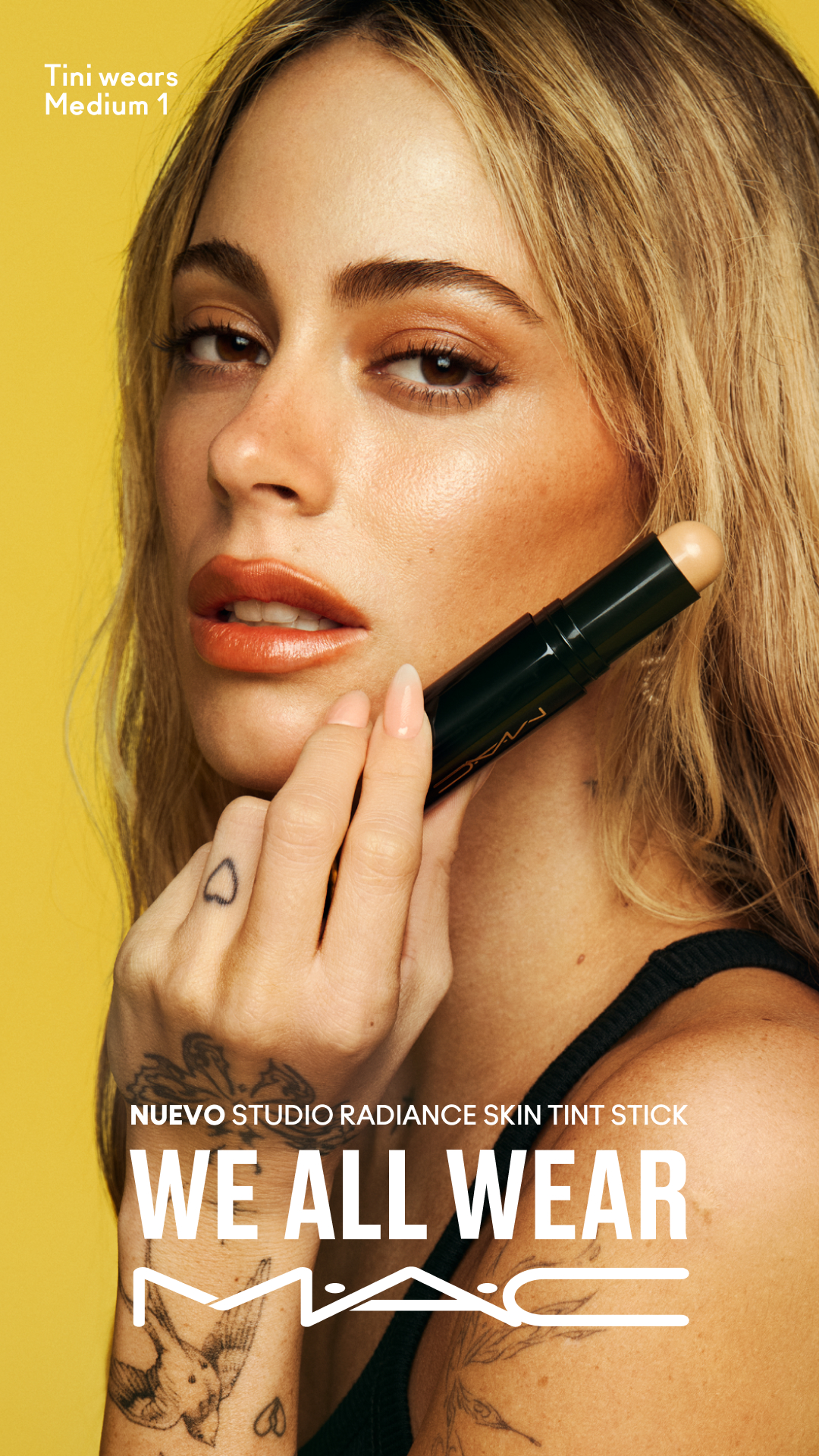 A close-up shot of a woman with blonde hair and makeup, holding a makeup stick near her face against a bright yellow background. Text overlay on the top left reads "Tini wears Medium 1". Below the woman's chin, larger text reads "NUEVO STUDIO RADIANCE SKIN TINT STICK WE ALL WEAR MAC" with the MAC logo at the bottom. The woman is looking towards the camera with a neutral expression, and the makeup stick is positioned near her lips.