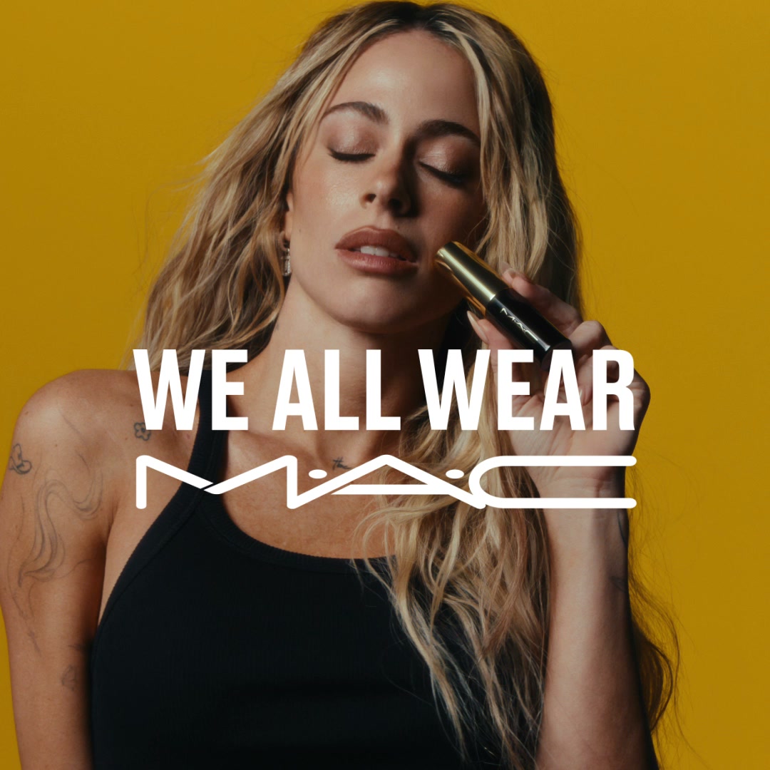 A woman with blonde hair and closed eyes is holding a MAC mascara wand near her face against a bright yellow background. Large white text overlays the image, reading "WE ALL WEAR" above the MAC logo. The woman is wearing a black tank top and has tattoos on her arm. The overall aesthetic is clean and focused on the brand and the product.