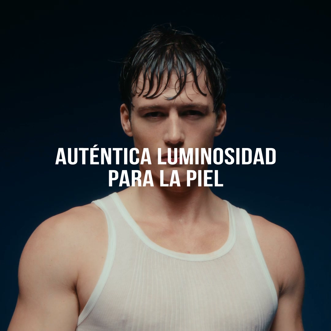 A close-up shot of a man with wet hair, wearing a white tank top, against a dark blue background. White text overlaid on the image reads "AUT uevoNTICA LUMINOSIDAD PARA LA PIEL" which translates to "Authentic luminosity for the skin." The man is looking directly at the camera with a serious expression. The overall aesthetic is clean and focused on the subject's skin and appearance.