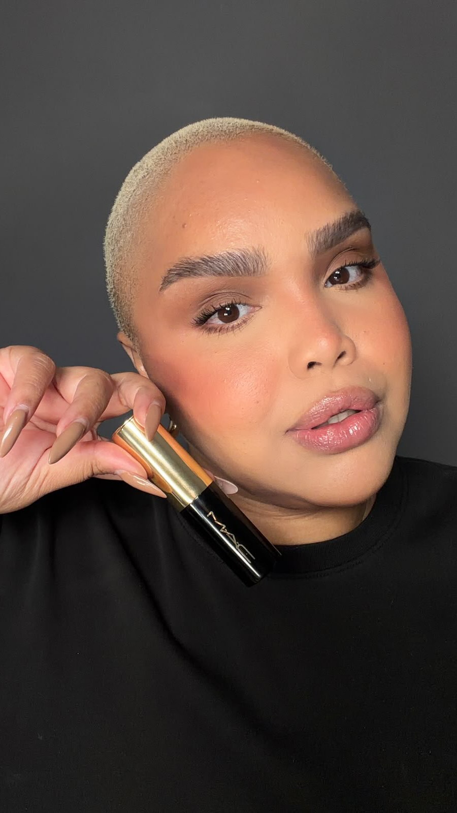 A close-up shot features a person with a shaved head and full makeup, holding a black and gold MAC cosmetic stick near their face. The product is angled to show the "MAC" logo. The background is a plain dark gray. On-screen text introduces the new Studio Radiance Skin Tint Stick from MAC Cosmetics, highlighting its hydrating, long-lasting properties and convenience. A discount code "MAC-ANGELICA" is provided for 10% off, with exclusions listed.