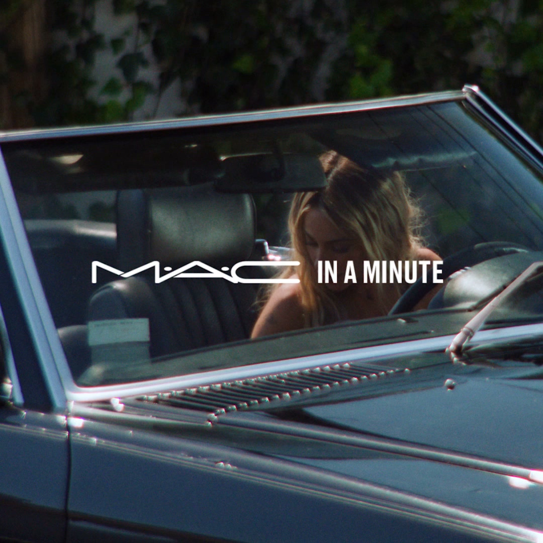 A woman with blonde hair is driving a convertible car. The MAC Cosmetics logo is overlaid on the windshield, along with the text "IN A MINUTE". The image is a lifestyle shot of someone in a car, with the brand prominently displayed. The text suggests a quick application or benefit related to the product.
