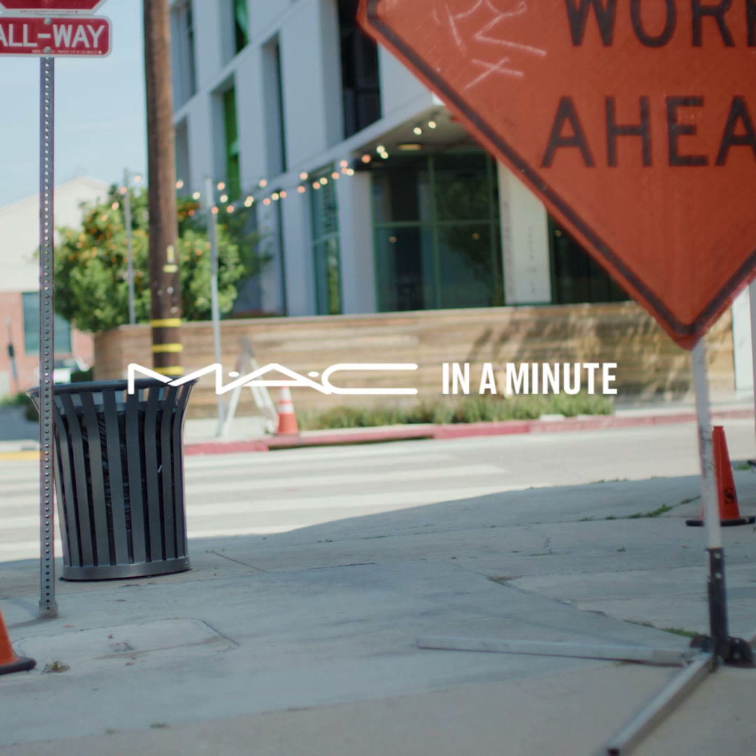 The image is an urban street scene with a "WORK AHEAD" sign in the foreground. The MAC Cosmetics logo and the text "IN A MINUTE" are overlaid in white, bold lettering across the middle of the frame. The background shows a modern building and trees. The overall impression is a blend of urban grit and brand presence, suggesting quick and easy application of MAC products in a busy environment.