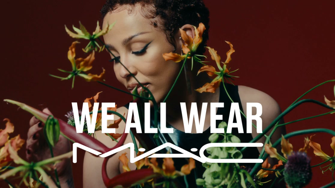A close-up shot of a woman with short, curly hair, looking down with her eyes closed. She is surrounded by vibrant orange and green flowers and foliage. Large white text overlays the image, reading "WE ALL WEAR" above the MAC Cosmetics logo. The overall aesthetic is artistic and emphasizes natural beauty and the brand's connection to it.