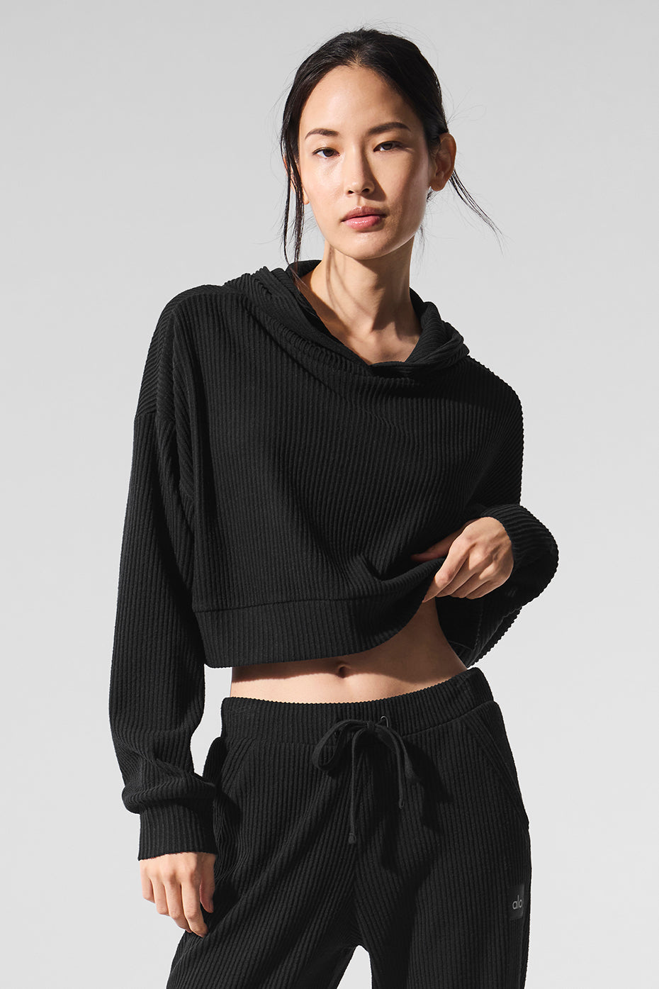 A woman with dark hair pulled back stands against a plain grey background, wearing a black ribbed cropped hoodie and matching sweatpants. She is pulling up the hem of her hoodie to reveal her midriff. The text "Closet refresh, incoming." is overlaid in white sans-serif font at the top of the image. The overall aesthetic suggests comfort and style, hinting at an update to one's wardrobe.