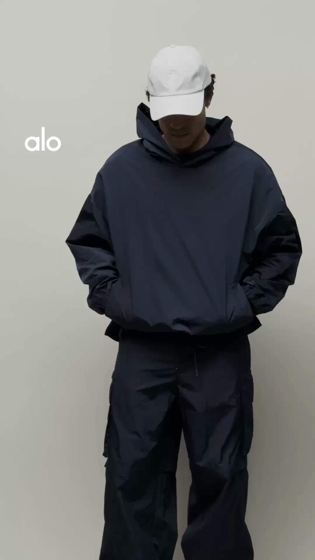 A person stands against a plain background wearing a dark blue hooded jacket and matching pants, with a white baseball cap on their head. The brand logo 'alo' is visible in white text on the left side of the image. Text overlaying the image reads: 'A classic shade that brings a sharp perspective to your everyday rotation.'