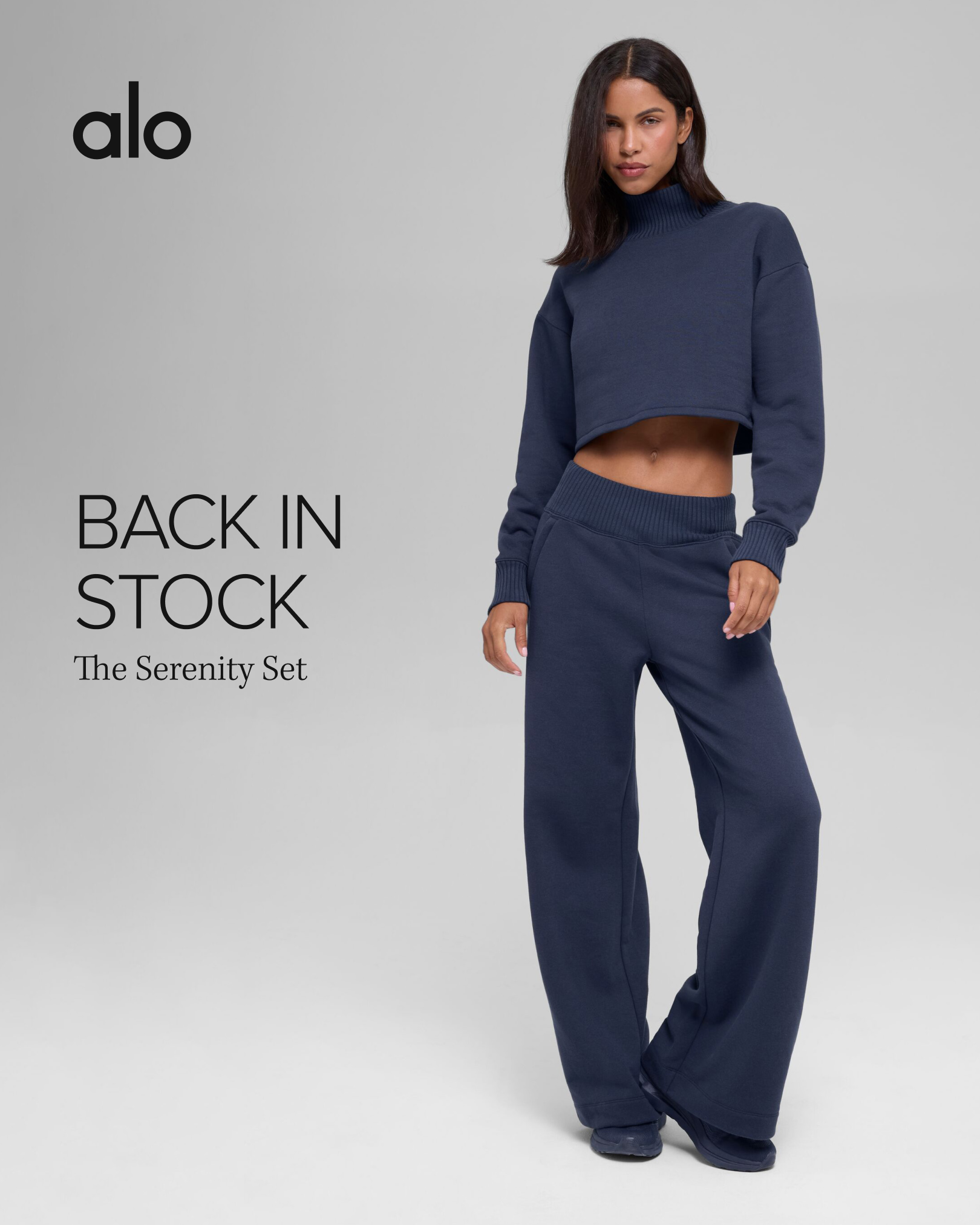 A female model stands against a plain grey background, wearing a navy blue matching set consisting of a cropped turtleneck sweatshirt and wide-leg sweatpants. The Alo Yoga logo is visible in the top left corner. Text on the left side of the image reads "BACK IN STOCK" in large, bold font, with "The Serenity Set" written below it in a smaller, elegant font. The creative highlights that a popular product is available again, implying limited availability.