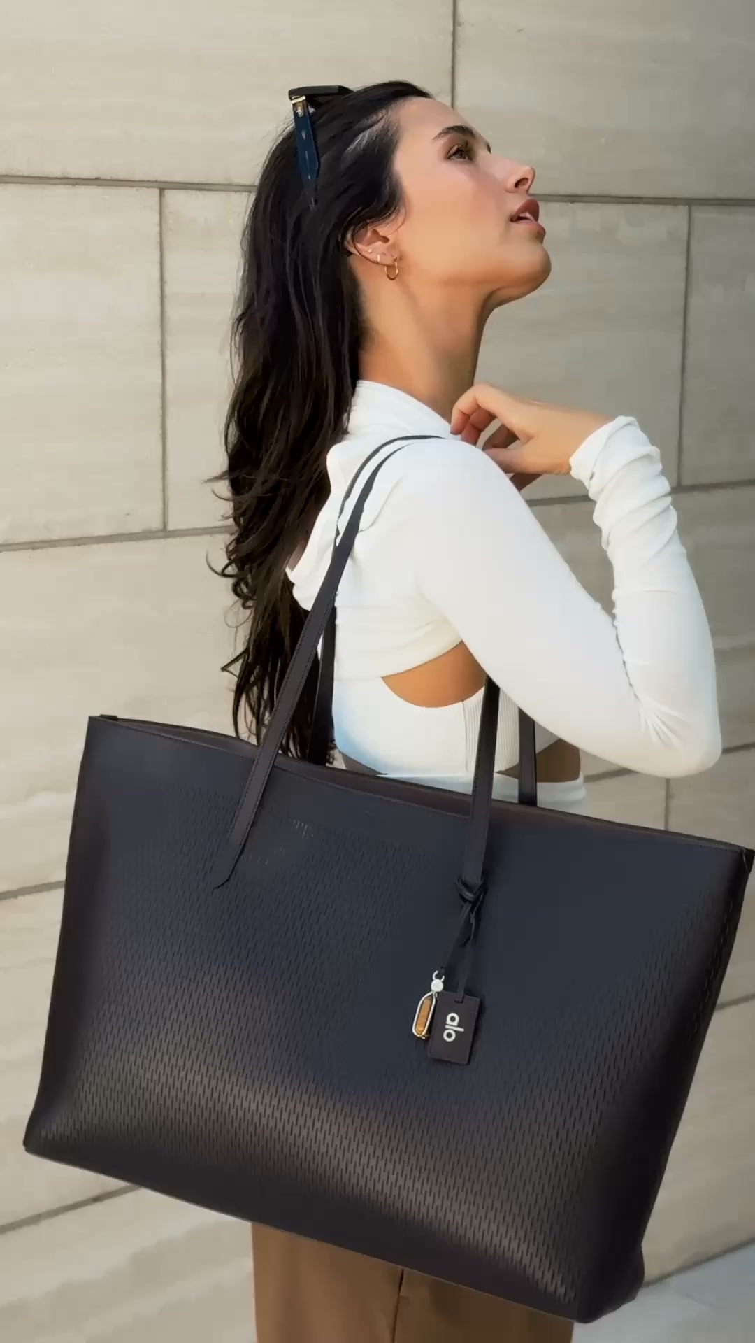 A woman with long dark hair, wearing a white cut-out top, is shown from the chest up, looking upwards and to the right. She is holding a large, dark brown tote bag with a perforated pattern. The bag has a small tag with the Alo Yoga logo. The background is a textured, light-colored wall. Text overlaying the image reads: "Playing favorites in the newest Java Brown drop."