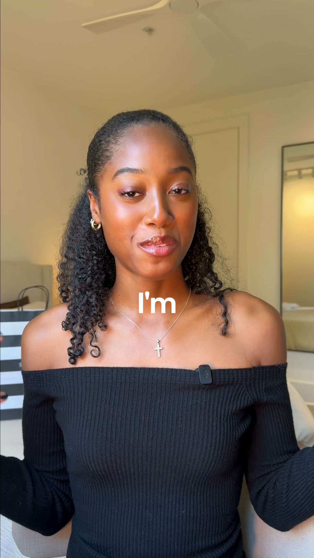 A young Black woman with curly hair pulled back is looking directly at the camera. She is wearing an off-the-shoulder black top and a silver necklace with a cross pendant. White text that reads "I'm" is overlaid on her chest. The background appears to be a softly lit room with a ceiling fan visible.