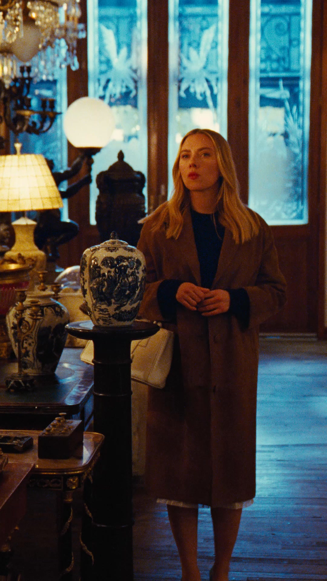 A blonde woman with red lipstick stands in an opulent room filled with antique furniture and decorative vases. She is wearing a brown coat over a dark sweater and a white skirt. The text overlay reads "Un simbolo di eleganza, senza tempo eppure in continua evoluzione." The ad features Scarlett Johansson and highlights the Prada Galleria bag.