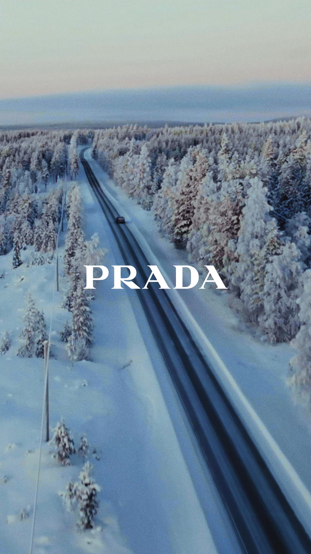 An aerial view shows a car driving on a long, straight road through a snow-covered forest. The trees are laden with snow, and the sky is a soft, pale blue. The word "PRADA" is displayed in large, white, serif font across the middle of the image. The copy describes a journey in a snowy landscape and a return, with the Holiday collection celebrating the season with poetic and informal elegance.