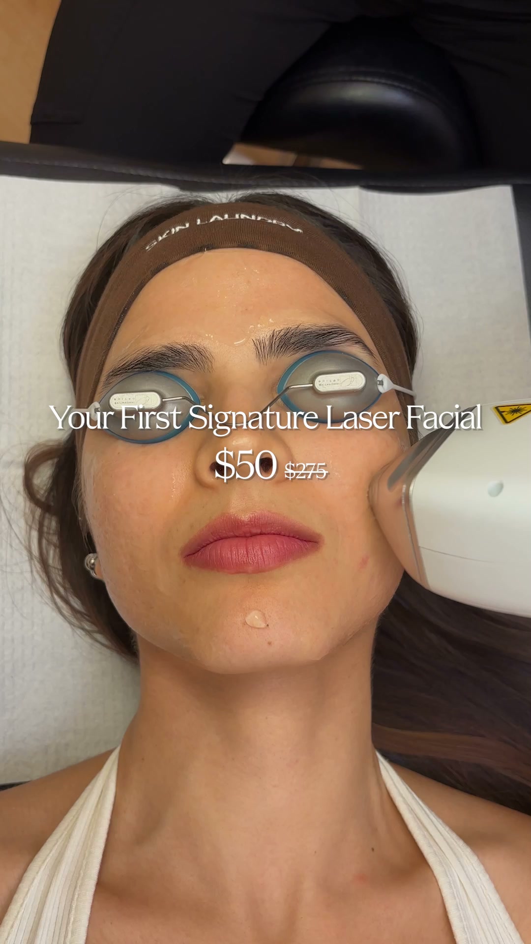 A close-up, overhead shot shows a woman lying down with protective goggles on her eyes and a brown headband on her forehead. A device is being held to her cheek, suggesting a cosmetic procedure. Text overlays the image, advertising "Your First Signature Laser Facial" for $50, with the original price of $275 crossed out, indicating a promotional offer.
