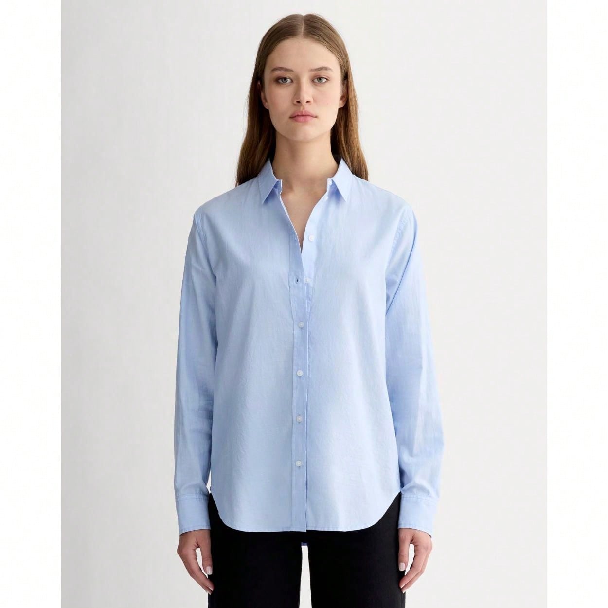A woman stands against a white background wearing a light blue button-down shirt and black pants. The image is a studio shot of a model. The text overlay reads: "From the cult-favorite tee to tissue tees, discover effortless basics you’ll love." The overall impression is one of simple, classic style.