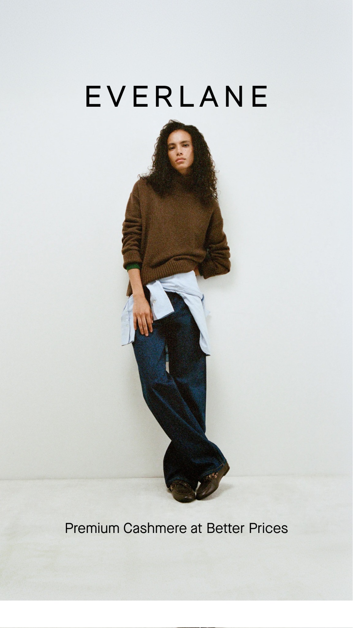A woman stands leaning against a white wall, wearing a brown sweater and dark jeans. The Everlane logo is displayed prominently at the top of the image. Below the woman, the text "Premium Cashmere at Better Prices" is overlaid on the image. The overall aesthetic is clean and minimalist, focusing on the product and brand.