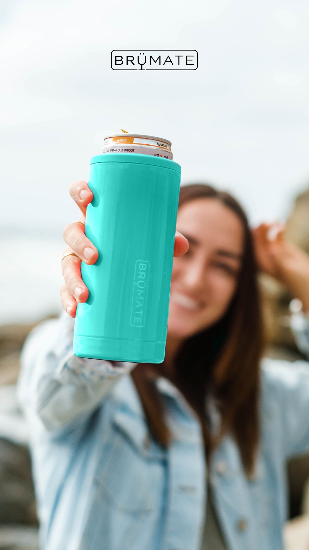 A woman is holding up a teal-colored Brumate can cooler to the camera, with a can inside. She is smiling and her face is slightly blurred. The background is a rocky beach with the ocean and sky visible. The Brumate logo is displayed at the top of the image. The entire text of a 5-star customer review from Danielle T. is overlaid on the image, praising the product's color and ability to keep drinks cold.