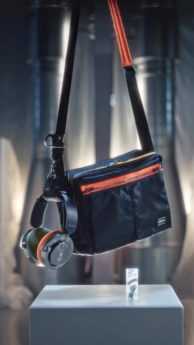 A black Porter bag with orange zippers hangs from a strap, with a pair of Dyson headphones also hanging from the strap. The bag and headphones are suspended above a white pedestal. In the background, large industrial pipes are visible. The text "Craftsmanship, encapsulated." is overlaid on the image.