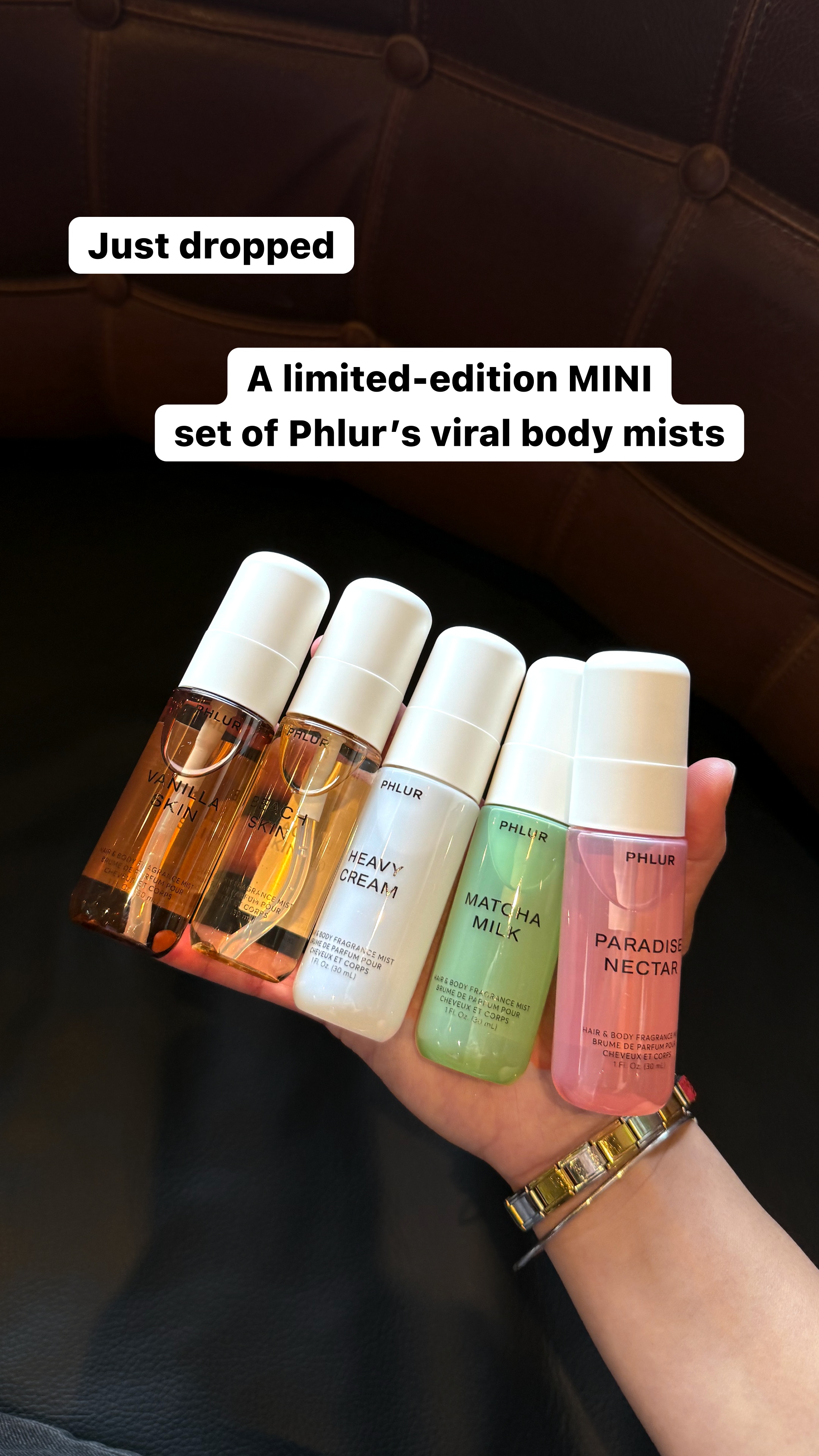 A hand holds five small bottles of Phlur body mists. The bottles are arranged in a slight curve, with the hand supporting them from below. The background is a dark, textured surface, possibly leather. White text overlays the image, with the main headline "Just dropped" in a rounded white box. Below it, in a larger font, it reads "A limited-edition MINI set of Phlur's viral body mists."