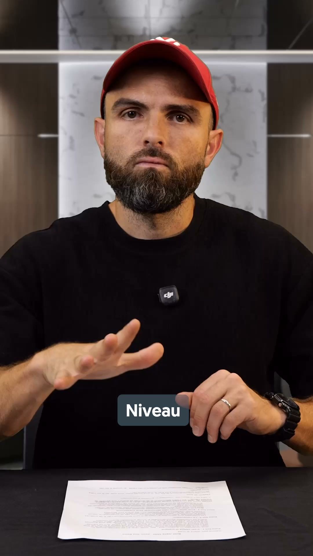 A man wearing a black t-shirt and a red baseball cap is looking directly at the camera. He has a beard and is wearing a ring on his left hand and a watch on his right. His left hand is raised, palm facing forward, and his right hand is resting on a table. A dark grey rectangular overlay with the word "Niveau" in white text is positioned in front of his right hand. A document is spread out on the table in front of him. The background appears to be an indoor setting with modern shelving or cabinetry.