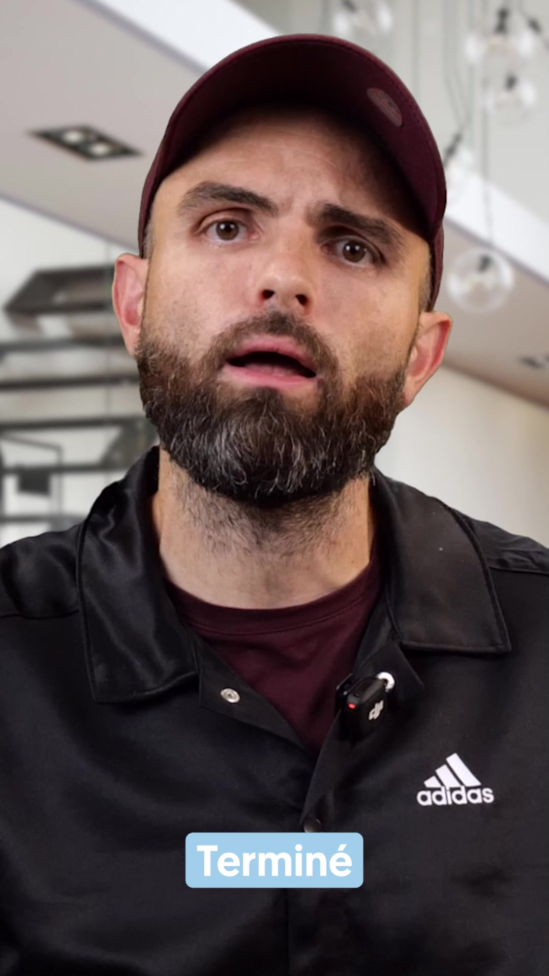 A close-up shot of a man wearing a maroon baseball cap and a black jacket with the Adidas logo. He has a beard and is looking directly at the camera with his mouth slightly open. A small DJI microphone is clipped to his jacket collar. A light blue rectangular text box with the word "Terminé" is overlaid on the lower part of the image. The background is blurred, showing what appears to be a modern interior with lights and shelving.
