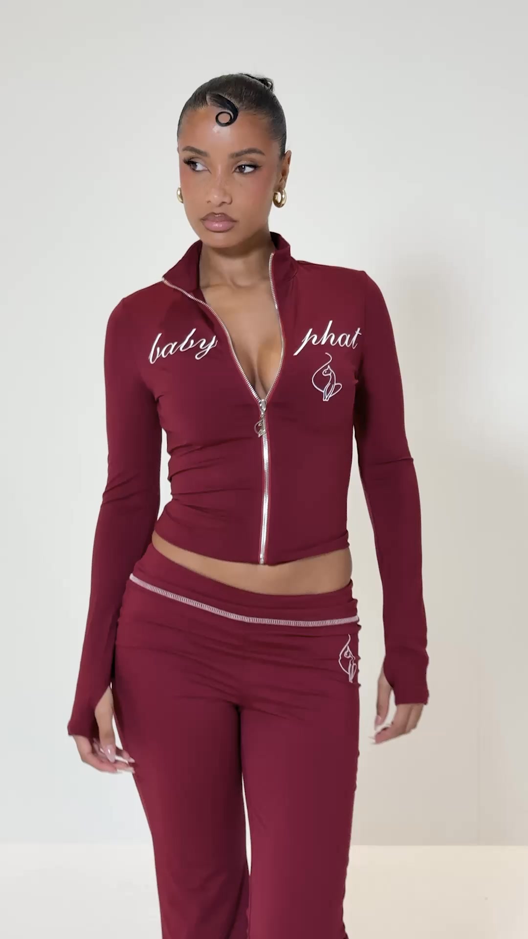 A young woman with dark hair styled in a distinctive swirl on her forehead stands against a plain white background. She is wearing a form-fitting, maroon-colored track jacket with a zipper partially undone, revealing a hint of cleavage, and matching pants. The jacket has the words "baby phat" embroidered in white script on the front, along with a small embroidered logo. The text "Matching sets. Maximum attitude. Baby Phat co-ords are back." is overlaid on the image.