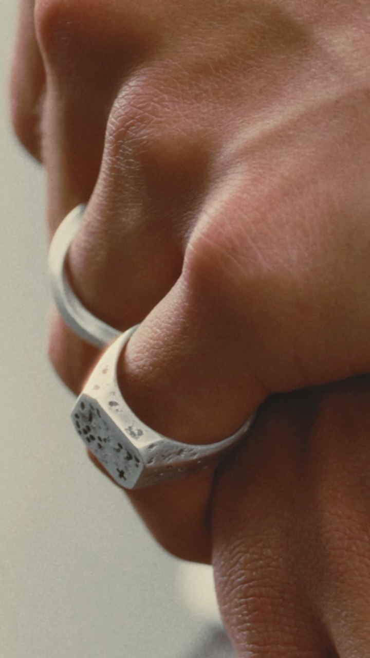 A close-up shot focuses on a hand wearing two silver rings. One ring is a signet ring with a textured surface and a patterned face, while the other is a simple band. The text "For the details that don’t go unnoticed." is overlaid on the image, followed by "Jewellery collection releases tomorrow." The image highlights the intricate details of the jewelry against the skin.