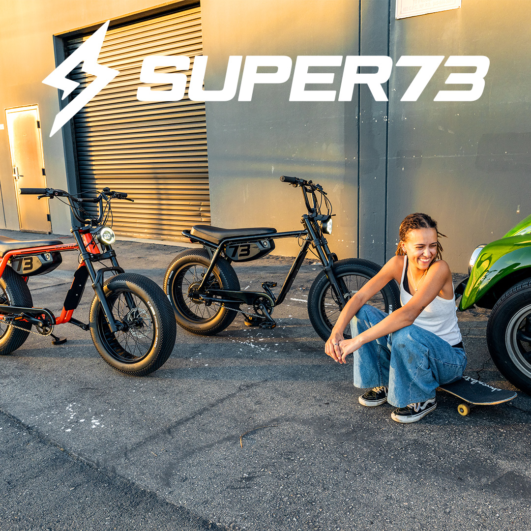 Two Super73 electric bikes are parked on a street. A young woman is sitting on a skateboard next to the bikes, laughing. The Super73 logo is prominently displayed in white text across the top of the image. The scene is set against a building with a large roll-up door and a bright, warm light illuminates the background, suggesting it might be sunset or sunrise. The overall image conveys a sense of freedom and urban adventure.