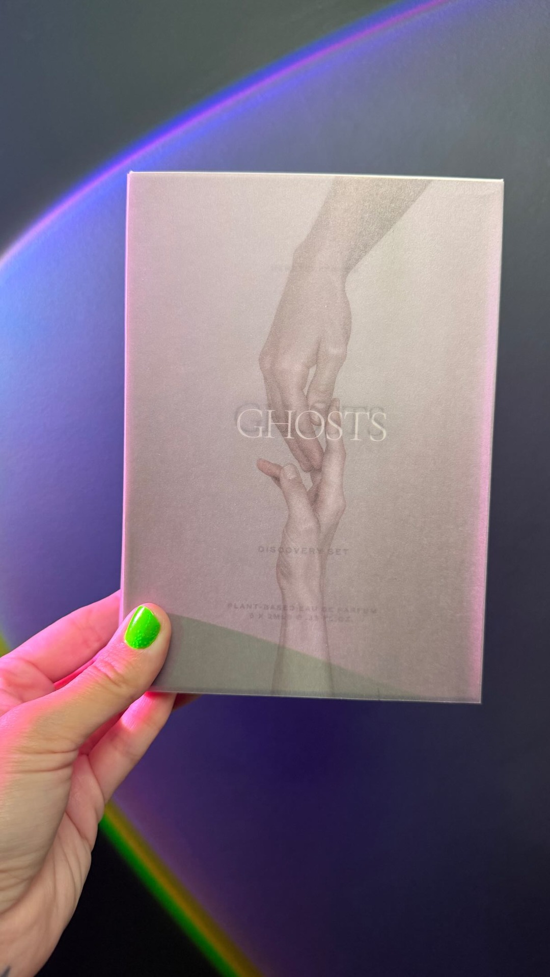 A hand with bright green nail polish holds a grey box with the word "GHOSTS" in large, elegant font. Below "GHOSTS" is smaller text that reads "DISCOVERY SET" and "PLANT-BASED EAU DE PARFUM". Two ethereal hands are depicted reaching for each other on the box. The background features a soft, blurred rainbow light effect. The text overlay asks the viewer to share their ghost stories.