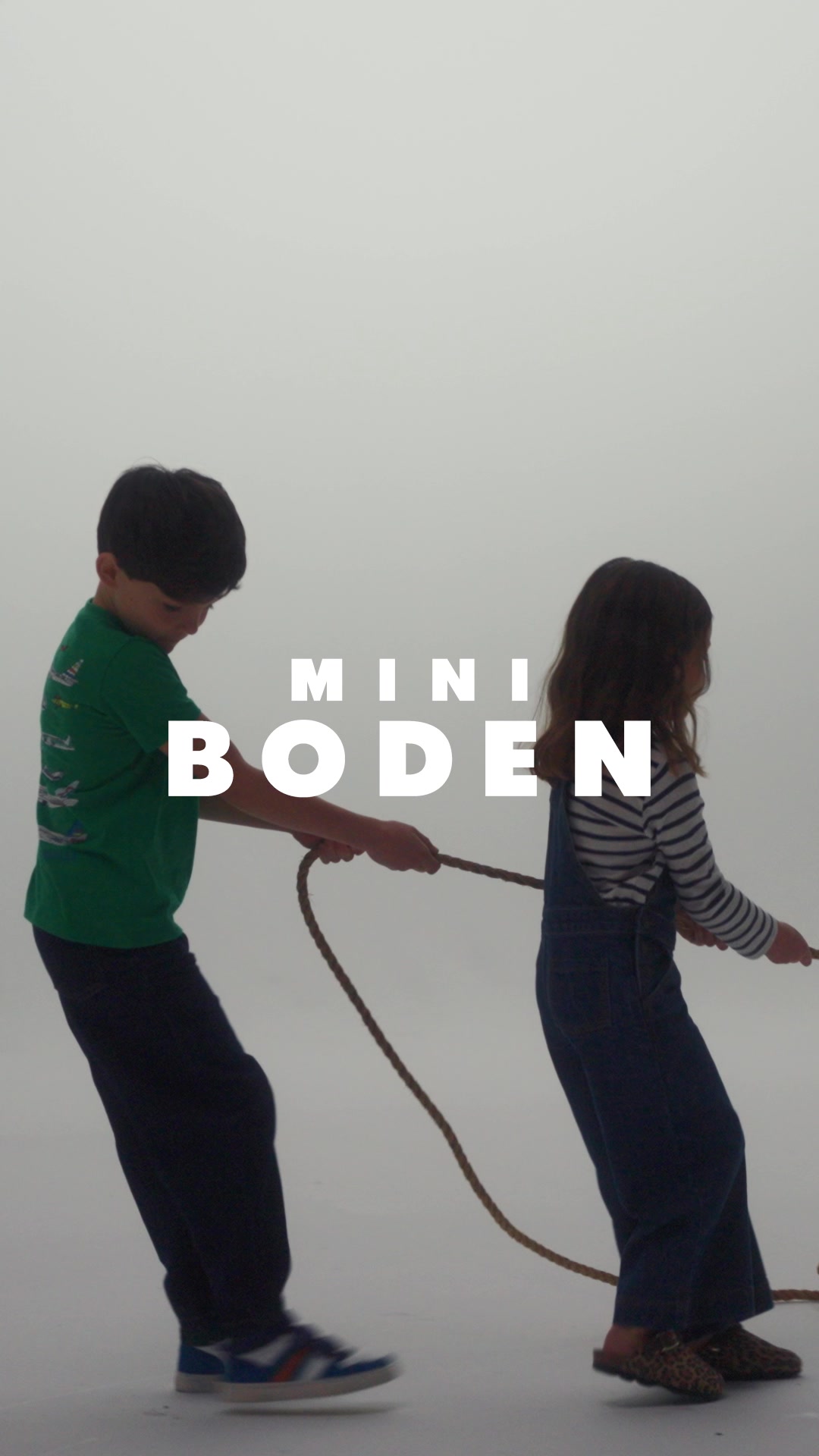 Two children, a boy and a girl, are playing tug-of-war with a rope. The text "MINI BODEN" is overlaid in large white letters across the center of the image. The boy wears a green t-shirt with graphics and dark pants, while the girl wears denim overalls over a striped long-sleeved shirt. The background is a plain, light gray, creating a studio-like setting. The creative appears to be explaining or introducing the "MINI BODEN" brand, possibly in relation to children's clothing or an activity book.