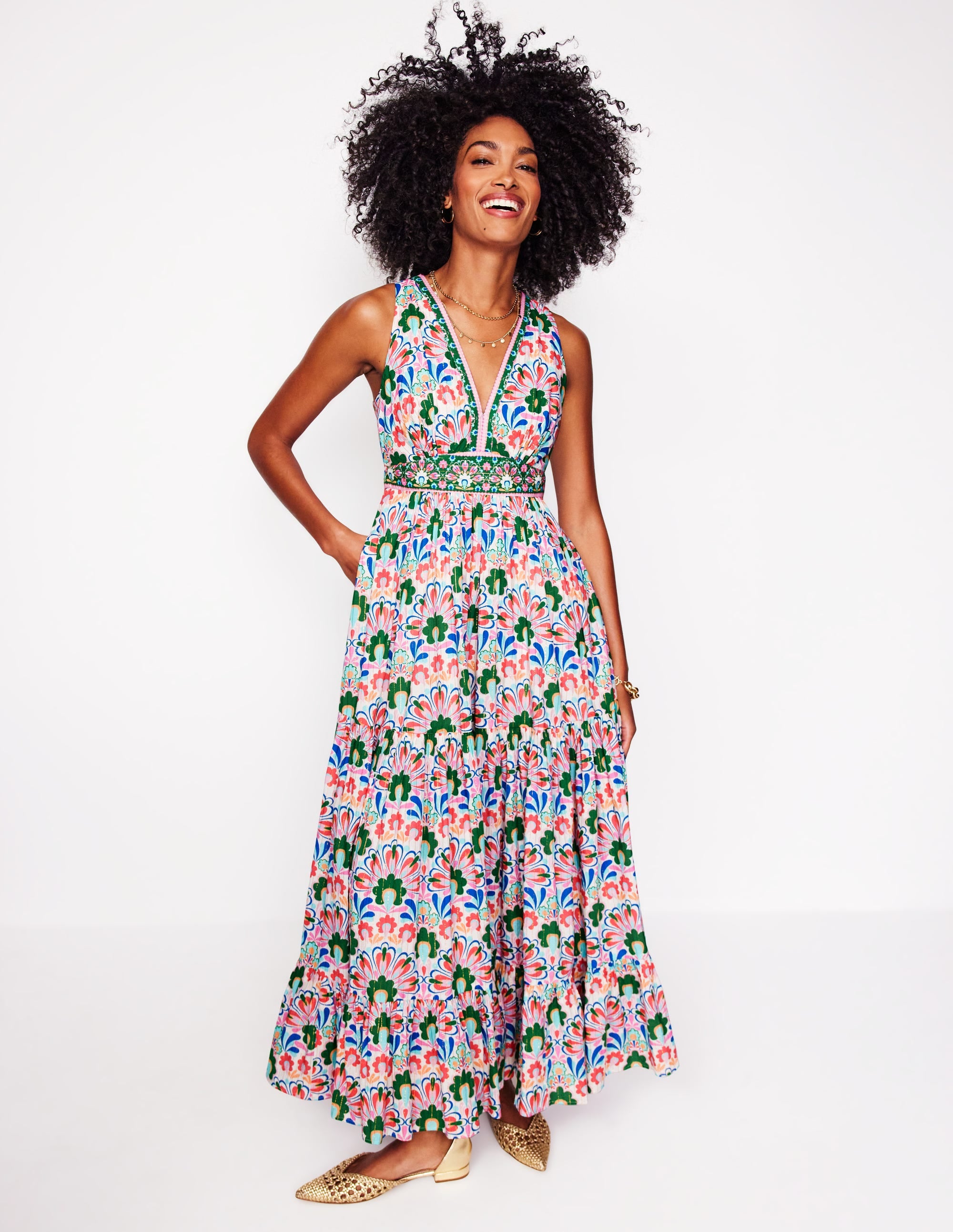 A woman with curly dark hair and a bright smile stands against a white background. She is wearing a colorful, floral-print maxi dress with a V-neck and a beaded belt. The dress has a tiered skirt and is sleeveless. She has her left hand in her pocket and her right arm is bent at the elbow. The text overlay reads: "Bei Boden wissen unsere Designer, ein oder zwei Dinge über mühelosen Stil. Wo auch immer Sie hingehen, unsere Kleider werden abliefern."