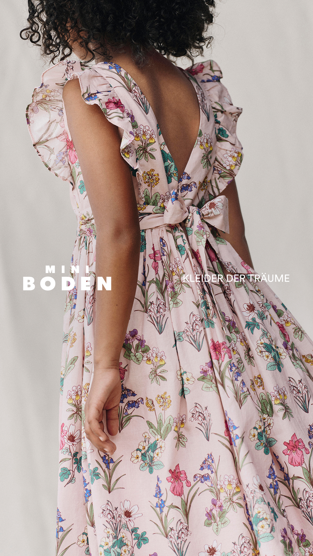 A young girl, seen from behind, is wearing a pink floral dress with ruffled sleeves and a bow tied at the back. The text "MINI BODEN" is overlaid on the left side of the image in large white sans-serif font, and "KLEIDER DER TRÄUME" (Dresses of Dreams) is overlaid on the right side in a smaller white font. The overall impression is one of whimsical childhood fashion.