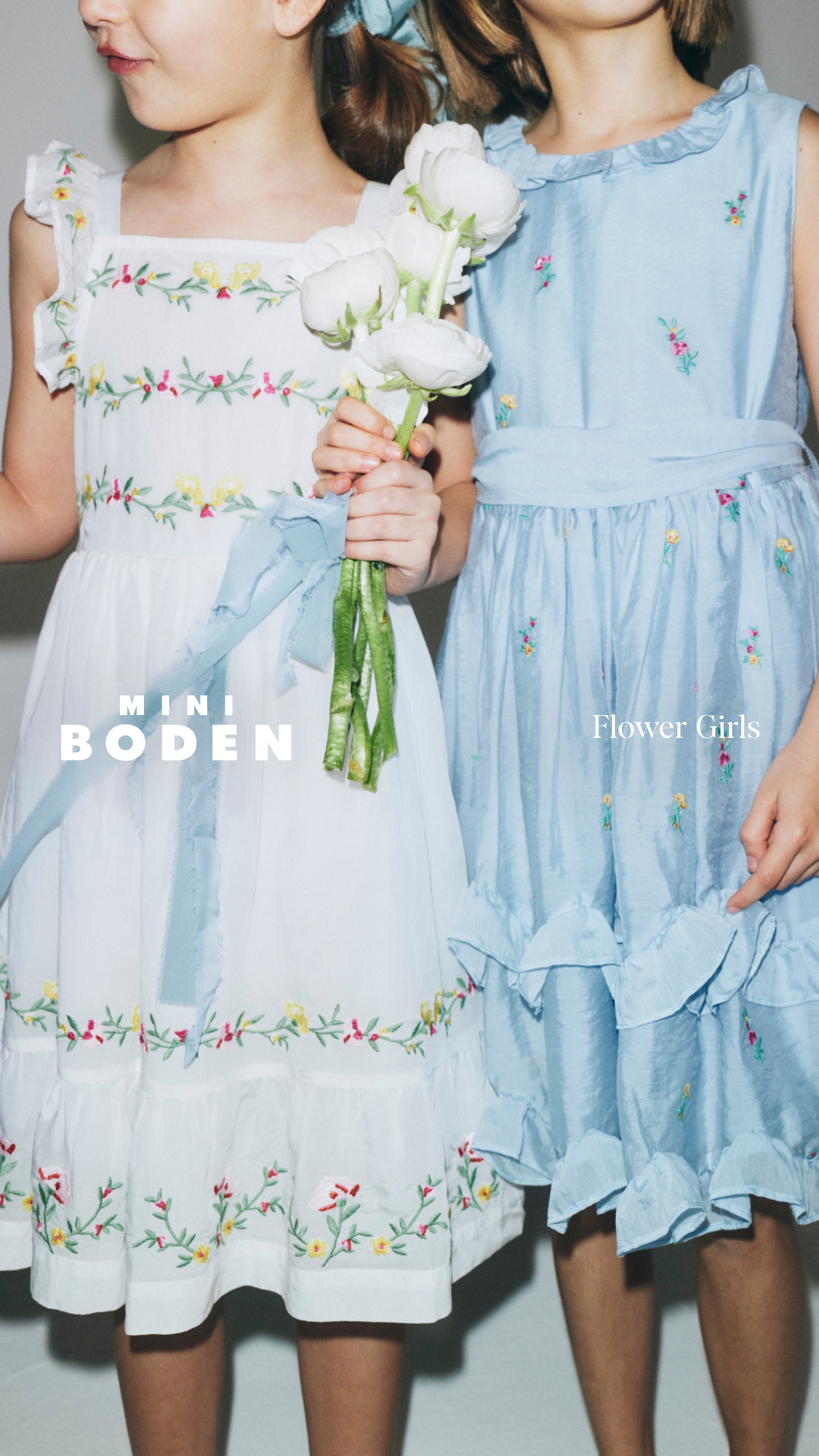 Two young girls are shown from the waist down, standing side-by-side. The girl on the left wears a white embroidered dress with yellow and pink flowers, holding a bouquet of white ranunculus flowers. The girl on the right wears a light blue embroidered dress with a ruffled neckline and hem. The text "MINI BODEN" is overlaid on the left side of the image, and "Flower Girls" is overlaid on the right side of the image.