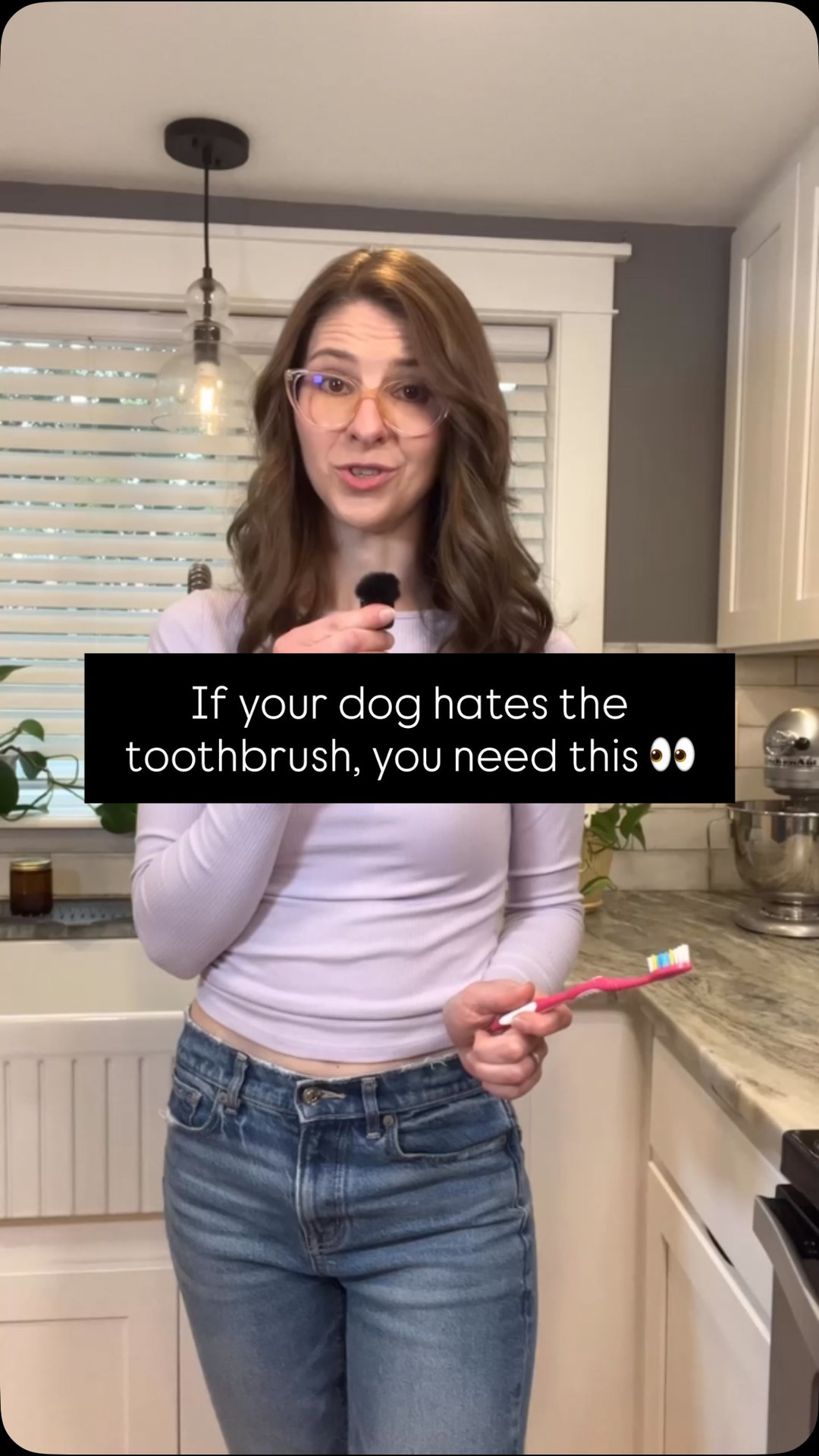 A woman stands in a kitchen holding a toothbrush. Text overlay on the image reads "If your dog hates the toothbrush, you need this 👀". The woman is wearing glasses and a light purple long-sleeved shirt with blue jeans. She is speaking into a microphone attached to her shirt. The background shows kitchen cabinets and a sink.