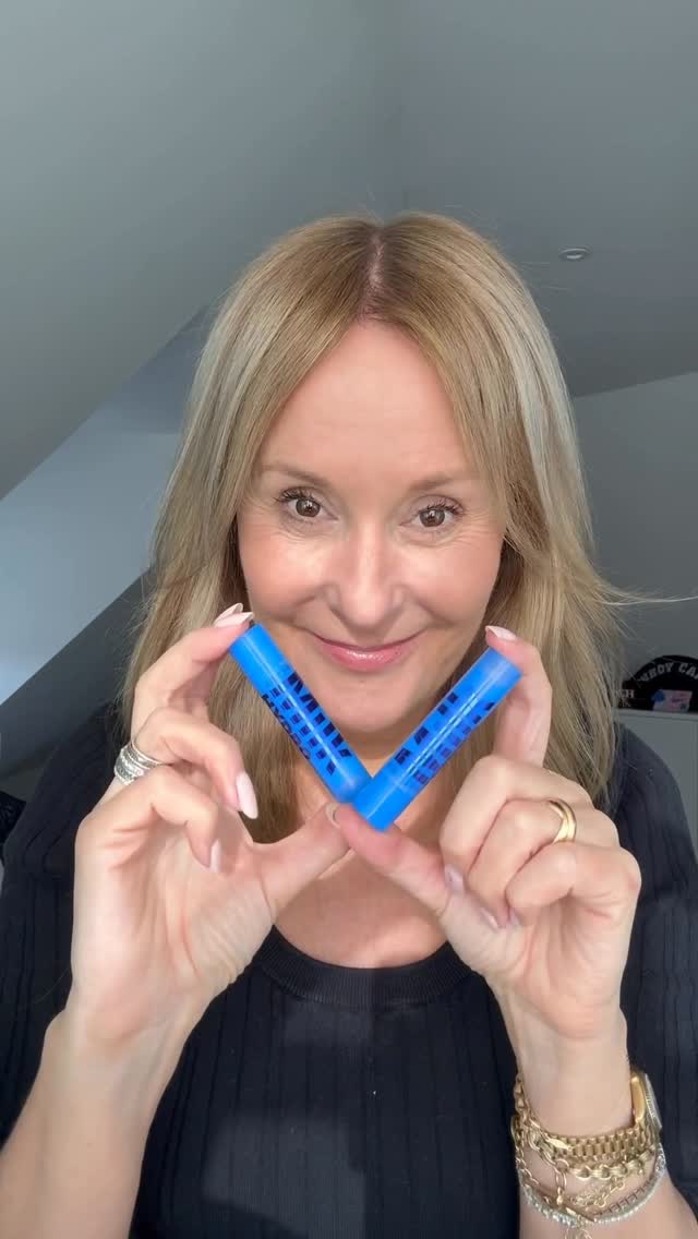 A woman with blonde hair and a black top holds up two blue tubes of Milk Makeup Hydro Grip Gel Concealer in a V-shape in front of her face. She is smiling and looking at the camera. Text overlaying the image reads "This Concealer is Genius". The copy describes the concealer as hydrating and beneficial for the skin, mentioning its coverage and longevity.