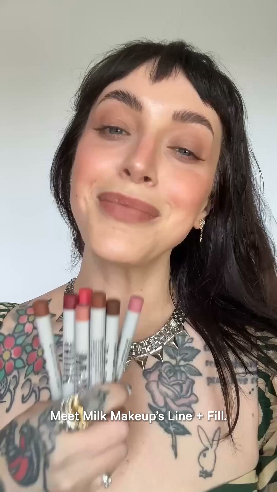 A woman with dark hair and bangs, wearing a spiked necklace, smiles at the camera and holds up several lip liners. She has tattoos on her chest and arms. On-screen text reads "Meet Milk Makeup's Line + Fill." The video appears to be a selfie-style recording from a creator showcasing the product.