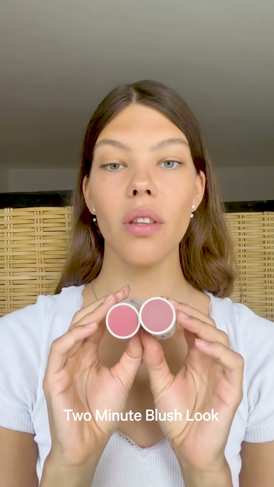 A young woman with brown hair and light makeup holds up two small tubes of blush, one pink and one a lighter shade of pink, in front of her face. The text "Two Minute Blush Look" is overlaid on the bottom of the image. The background is a woven bamboo screen. The creative appears to be demonstrating a quick makeup application technique.
