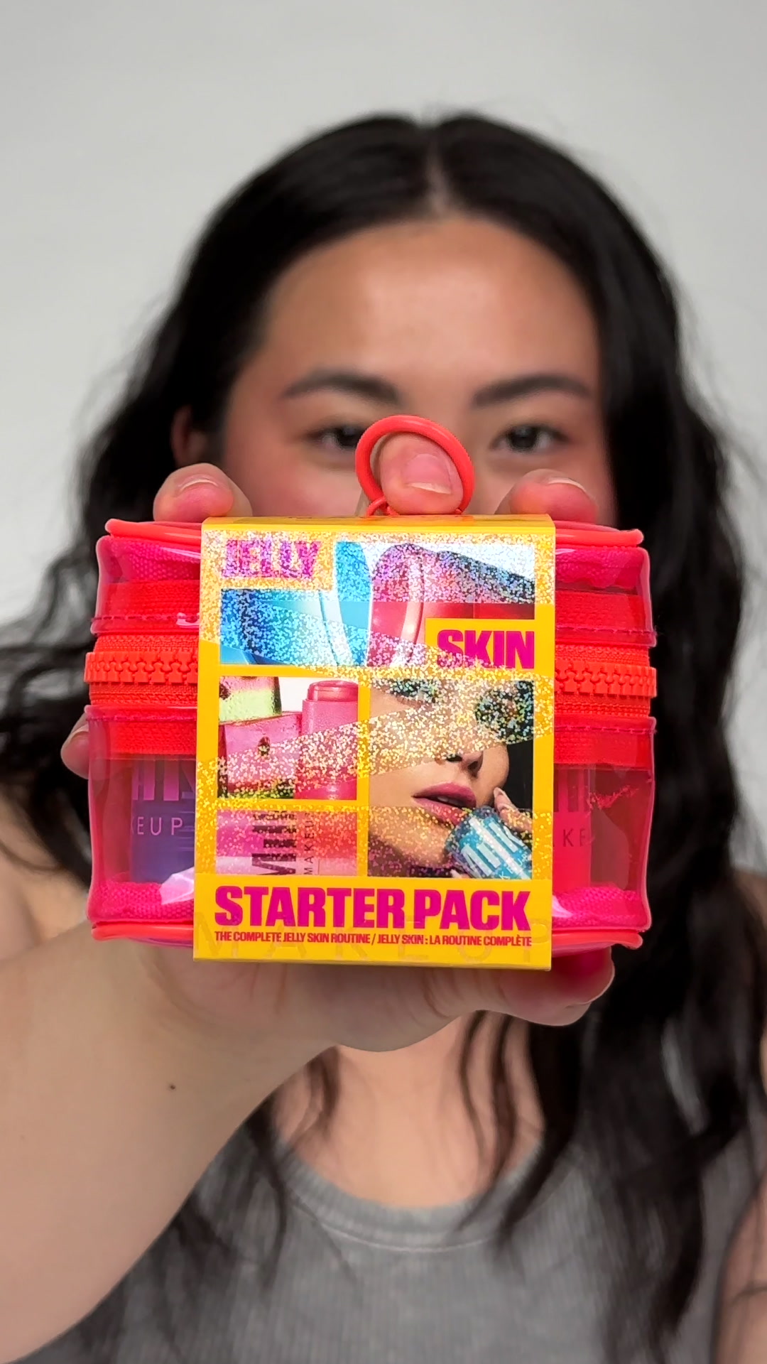 A person holds up a bright pink makeup bag containing a "Jelly Skin Starter Pack". The packaging is yellow and features a collage of images related to skincare and makeup, including a close-up of a woman's face with glitter. The text "JELLY SKIN STARTER PACK" is prominently displayed in bold, colorful letters. The copy, "Apaise. Illumine. Teinte. Notre Jelly Skin Starter Pack en édition limitée est là. Et c’est une offre qui ne rigole pas. Mets la main dessus avant qu'il ne disparaisse.", emphasizes the limited availability of the product.