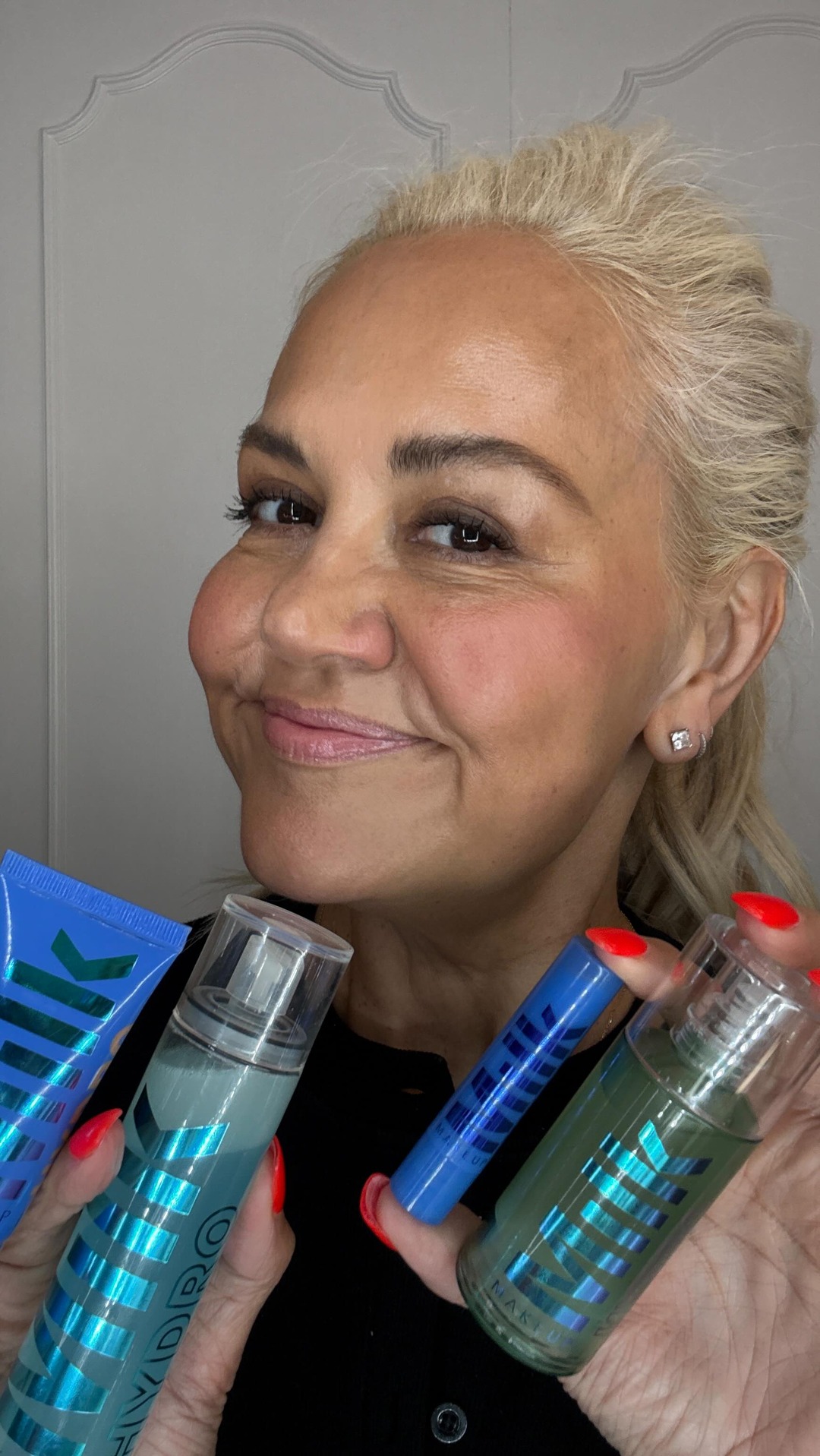 A woman with blonde hair pulled back smiles at the camera, holding up several Milk Makeup products. She is showcasing a blue tube, a clear spray bottle with blue liquid, a blue stick, and a green spray bottle. The background is a plain, light-colored wall with decorative molding at the top. The text overlay highlights the brand and the product collection, posing a question about achieving both hydration and longevity in makeup.
