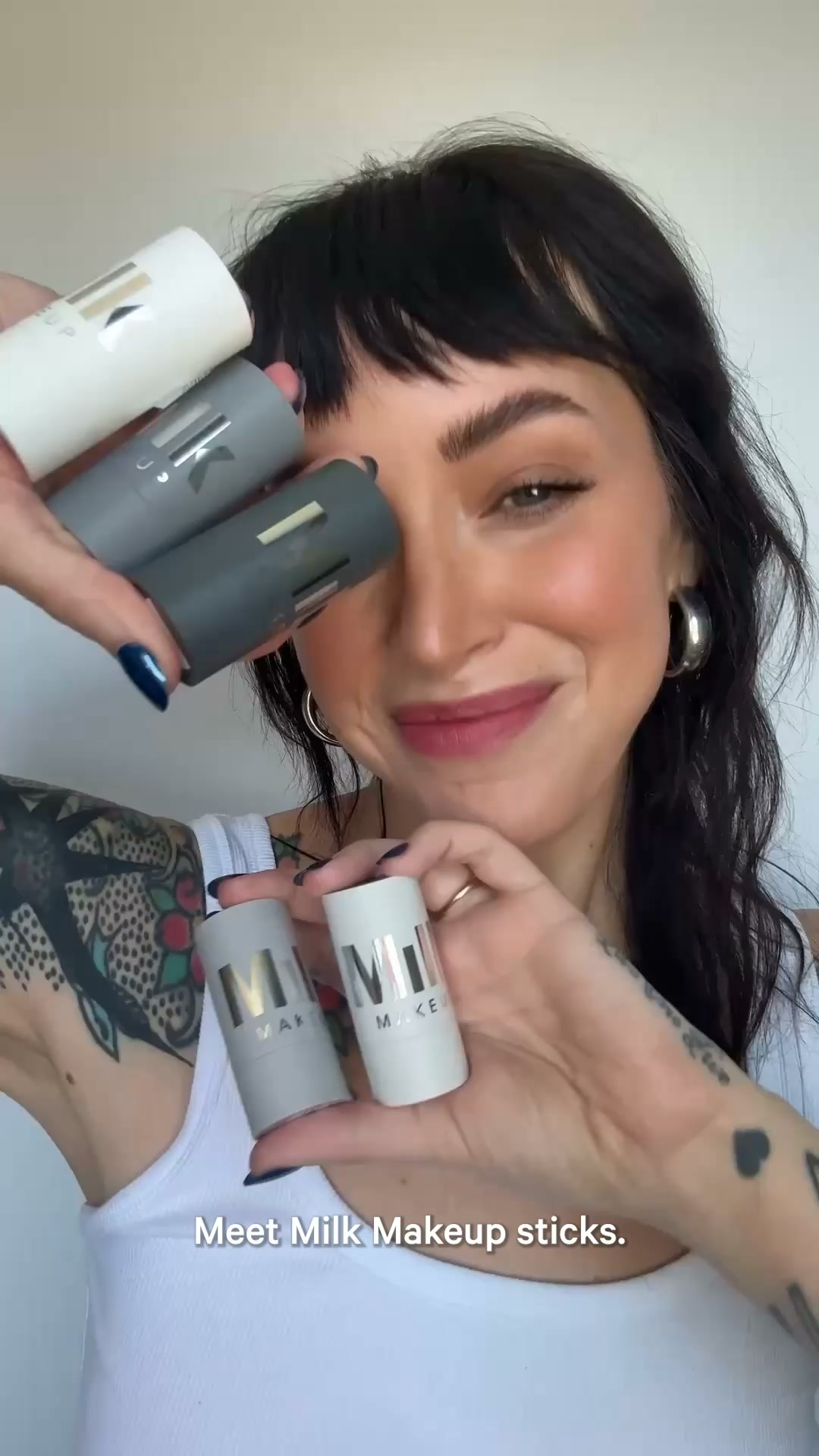 A woman with dark hair and bangs, wearing a white tank top, smiles as she holds up several Milk Makeup sticks. She has tattoos on her arm and is wearing large hoop earrings. The text "Meet Milk Makeup sticks." is overlaid on the bottom of the image. The background is a plain, light-colored wall.