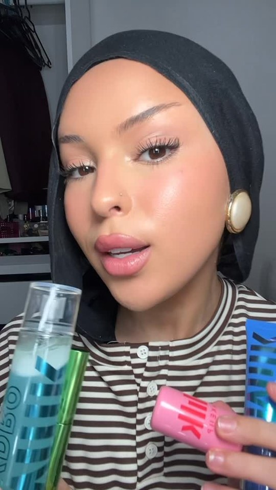 A woman wearing a black hijab and a striped shirt holds up several Milk Makeup products. She is looking directly at the camera with a slight smile. The products visible include a blue spray bottle, a green tube, and a pink stick. Text overlays the image, listing the products and where to find them. The overall impression is a personal recommendation from a creator.