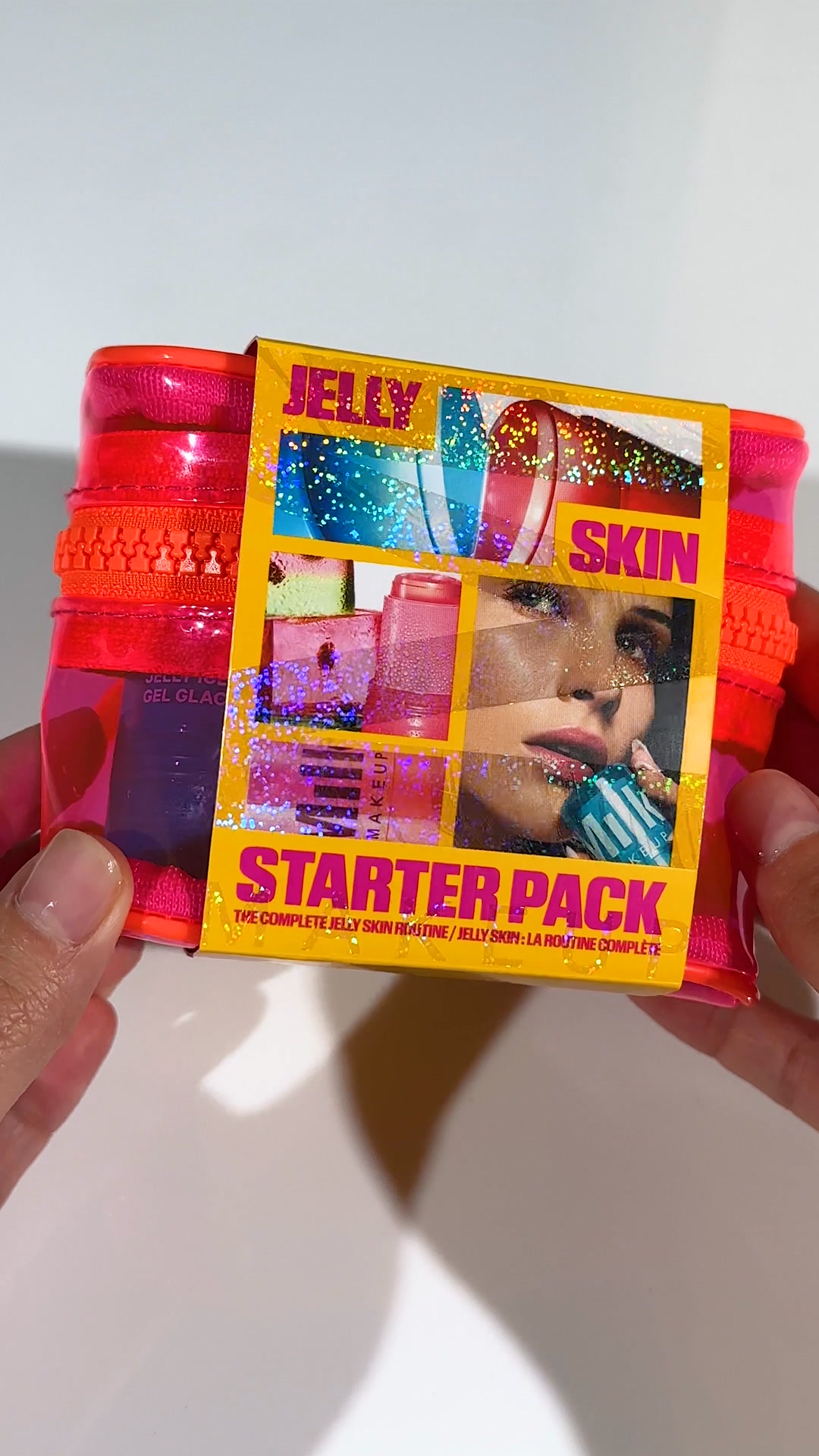 A bright pink cosmetic bag is held by two hands. A yellow label with glitter overlays the front of the bag, featuring a collage of product images and a close-up of a woman's face. The text on the label reads "JELLY SKIN STARTER PACK" and "THE COMPLETE JELLY SKIN ROUTINE / JELLY SKIN : LA ROUTINE COMPLÈTE". The overall aesthetic is vibrant and sparkly, suggesting a fun and effective skincare product.