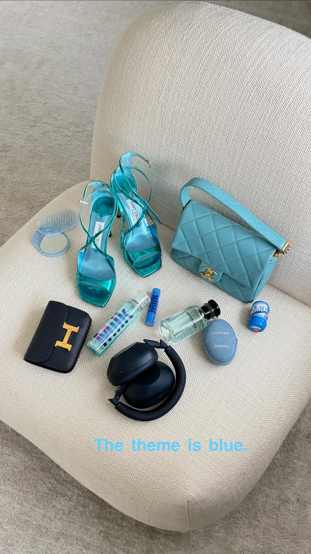 A flatlay of various blue and teal luxury items is arranged on a textured cream chair. The items include teal strappy heels, a quilted teal handbag, a navy wallet with a gold 'H' logo, a blue makeup compact, a blue lipstick or balm, a bottle of perfume, a spray bottle, and black headphones. The text "The theme is blue." is overlaid in a light blue, slightly transparent font across the bottom of the image.
