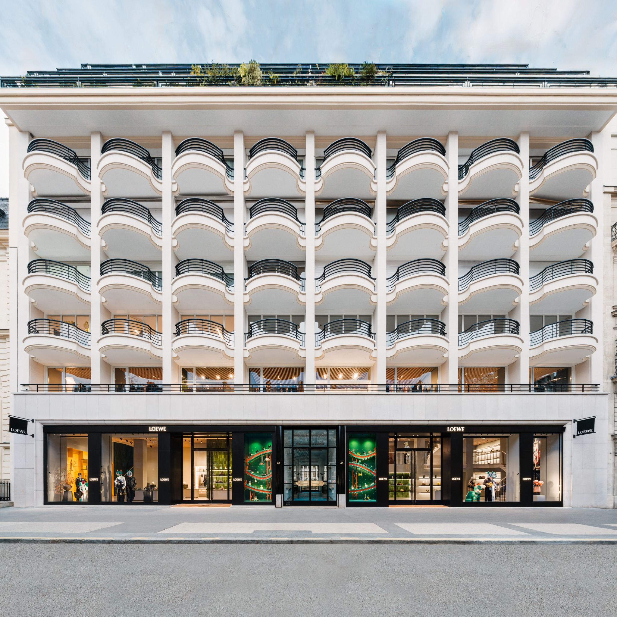 The image shows the exterior of a Loewe store with a modern, multi-story facade featuring curved balconies. The ground floor has large glass windows displaying store interiors with mannequins and products. The balconies above are white with black railings, and the top of the building has greenery. The overall impression is one of luxury and architectural design, showcasing the brand's physical presence.