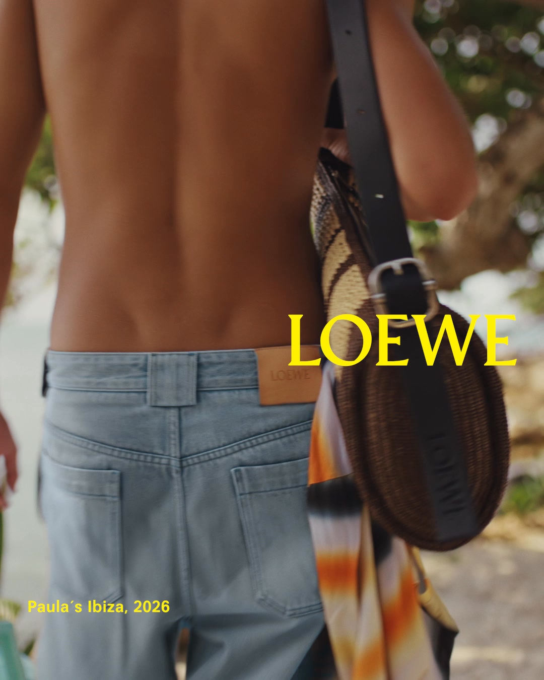 The back of a person wearing light blue denim shorts and a black bag slung over their shoulder is shown. The Loewe logo is prominently displayed in yellow text on the right side of the image. Below the person's shorts, the text 'Paula's Ibiza, 2026' is visible in yellow. The scene appears to be outdoors, possibly a beach or tropical location, with blurred foliage in the background.