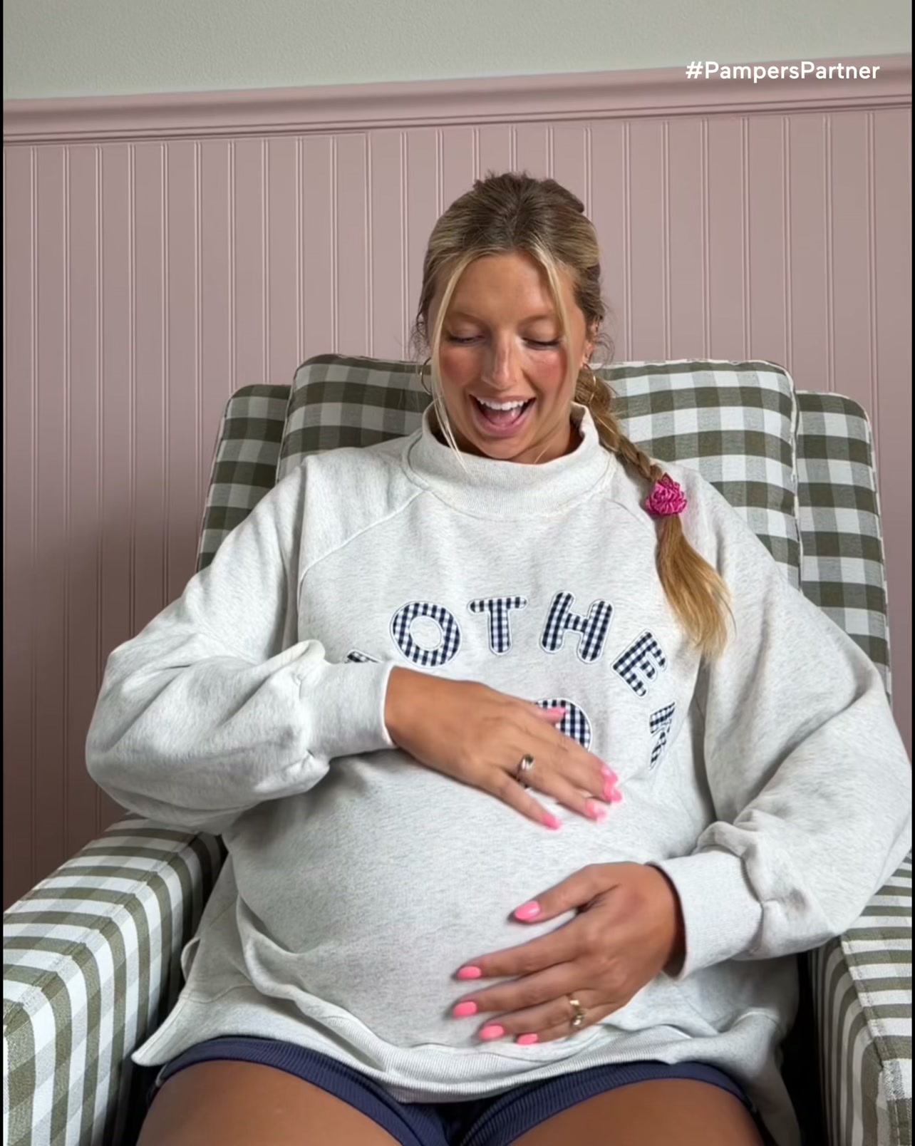 A pregnant woman with blonde hair in a braid, wearing a grey sweatshirt, sits in a chair and cradles her baby bump. She is smiling and looking down at her belly. The background features a pink paneled wall with a green and white checkered chair. The text "#PampersPartner" is visible in the upper right corner of the image.