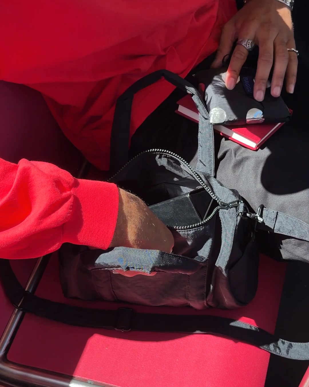 A close-up shot shows a person's hands reaching into an open black Baggu bag. The bag contains a small red notebook and a black wallet. The person is wearing a bright red long-sleeved shirt, and another person is wearing black pants and a red top. The background is a red surface, possibly a seat. Text overlays the image, announcing new arrivals and available patterns.