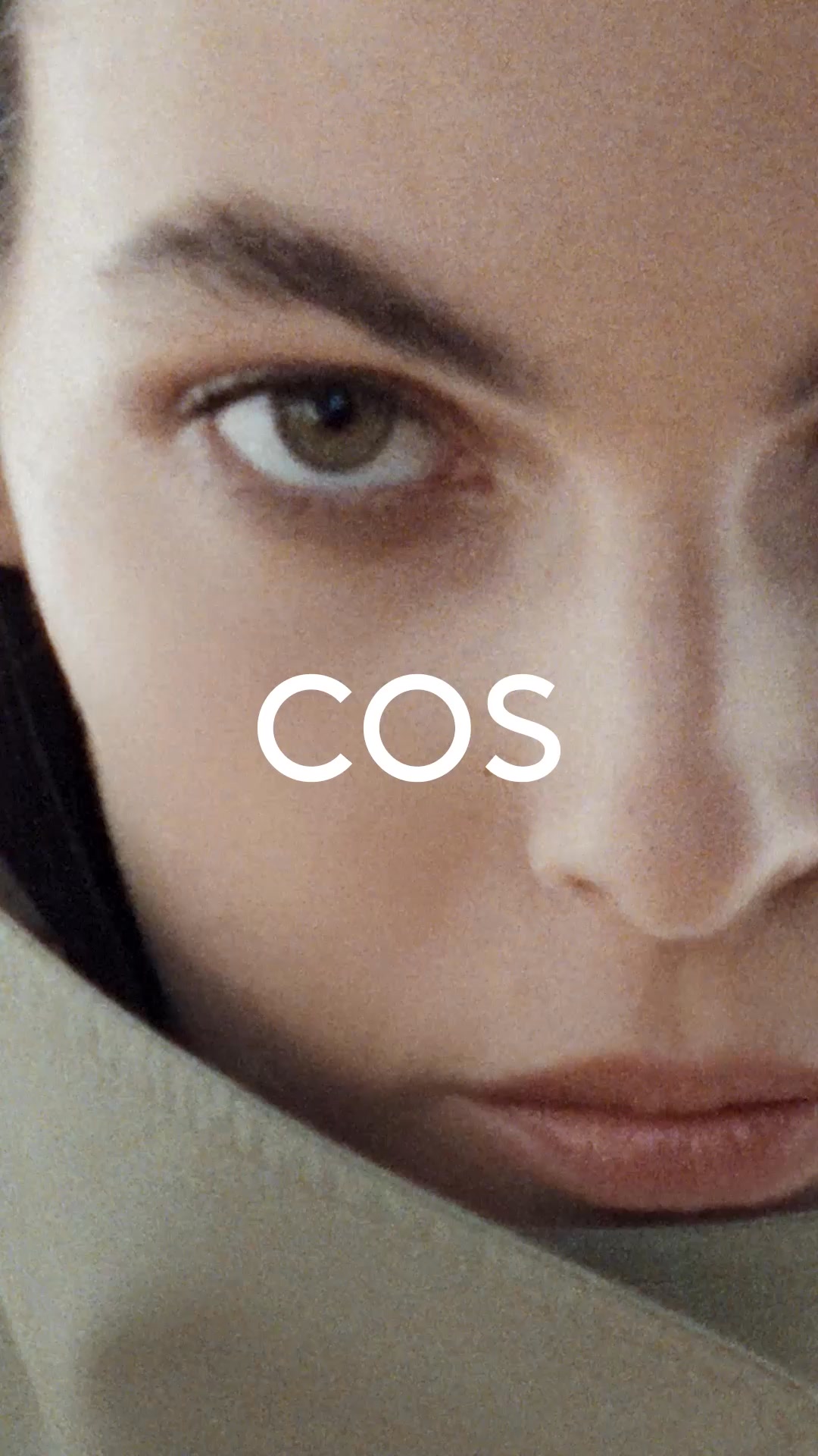 A close-up shot of a person's face, focusing on their eye and lips. The word "COS" is displayed in large white sans-serif font across the center of the image. The person is wearing a light-colored garment that partially covers their neck and shoulder. The overall aesthetic is minimalist and fashion-forward, with soft lighting and a shallow depth of field.